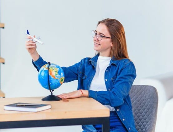 A young woman in glasses, sitting at a desk, holding a toy airplane and looking at a globe, symbolizing travel planning or tourism. 