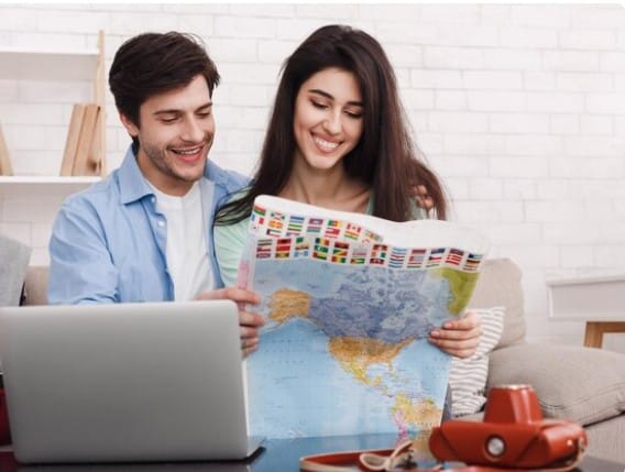 Smiling couple planning a trip, looking at a world map and using a laptop for research. Travel planning concept.