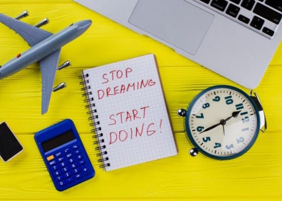 Stop Dreaming, Start Doing!' surrounded by a model airplane, calculator, alarm clock, and a laptop. Travel and goal-setting inspiration."